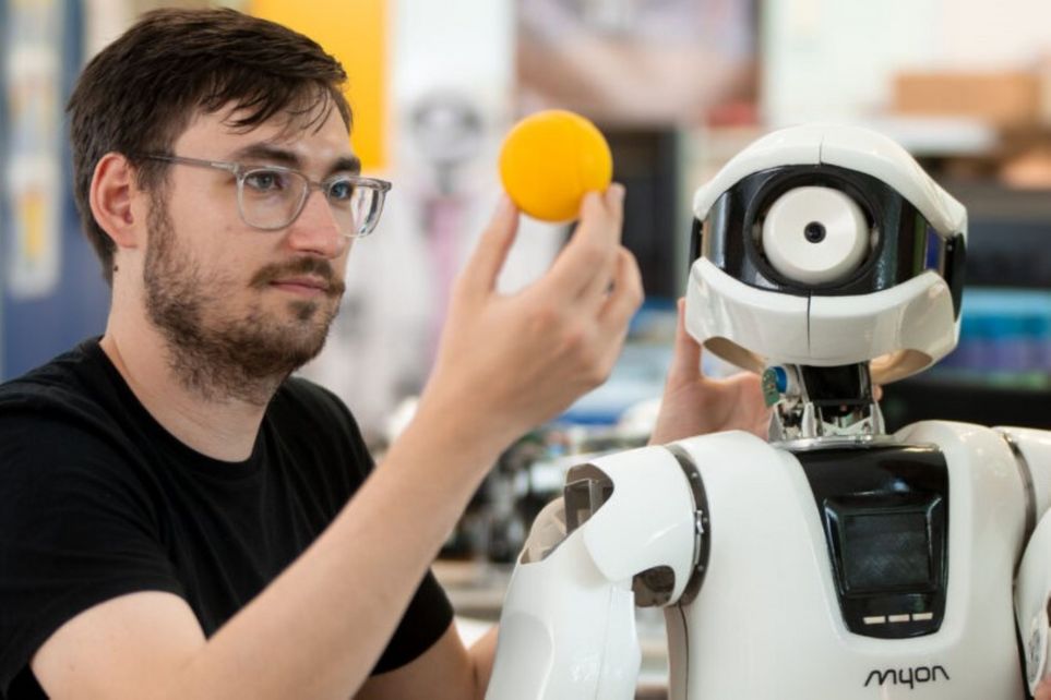 Ein Mensch hält einen gelben Ball in der Hand. Neben ihm ein humanoider Roboter, der auf den Ball schaut.