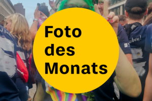 Bildaufschrift: „Foto des Monats“. Im Hintergrund ist eine Menschenmenge zu sehen.