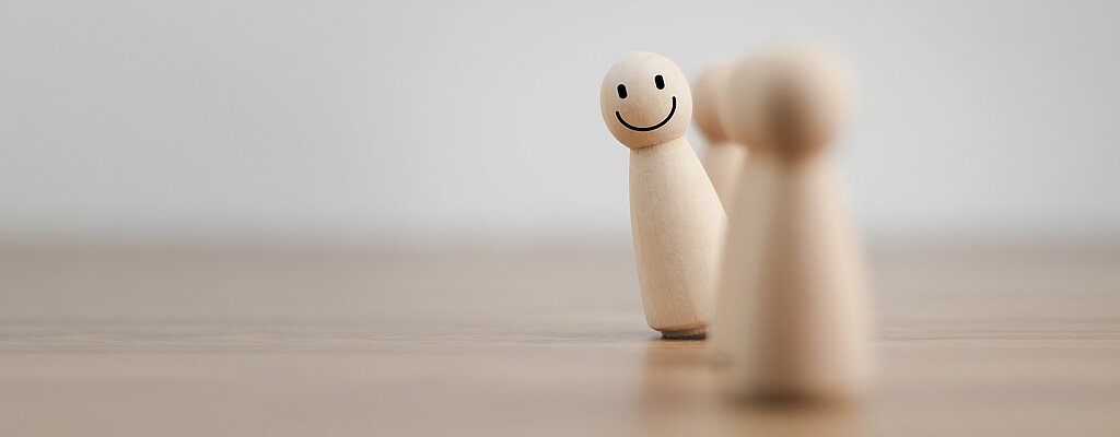 Ansprechpartner*innen Zwei Spielfiguren aus Holz auf einer Holzunterlage. Eine Figur hat ein lächelndes Gesicht aufgemalt