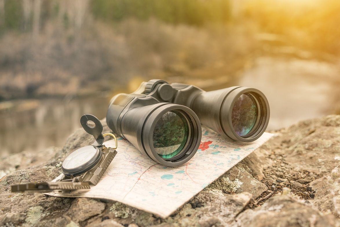 Fernglas und Kompass liegen auf einem Stein, dahinter ein Fluss und Wälder