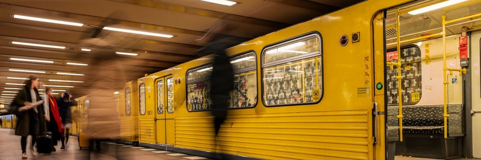 Eine gelbe U-Bahn steht am Gleis in einem U-Bahnhof. An ihr gehen Menschen vorbei, sie sind teilweise verschwommen.