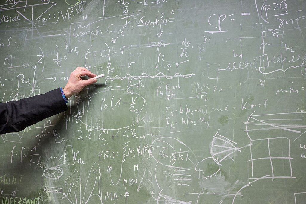 Labor für Numerische Mathematik. Foto einer mit Formeln vollgeschriebenen Tafel. Von links ragt der Arm eines Menschen hinein, der ein Stück Kreide in der Hand hält.