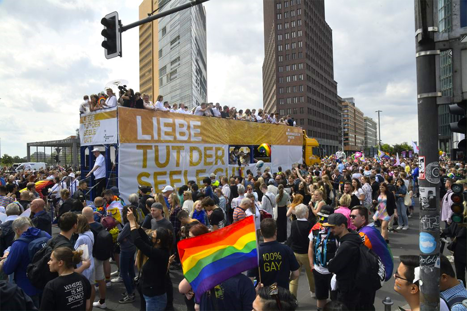 Demonstrationszug in der Innenstadt von Berlin: Eine Person schwenkt eine Regenbogenflagge, ein Bus trägt die Aufschrift „Liebe tut der Seele gut“.