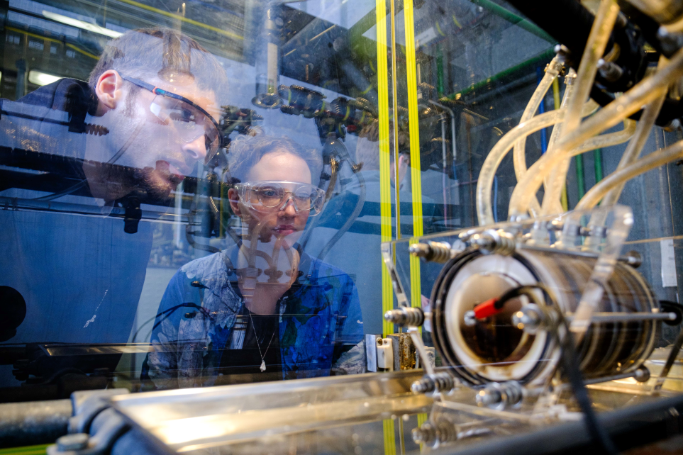 Ein Student und eine Studentin betrachten einen technischen Gegenstand durch eine Glasscheibe hindurch.