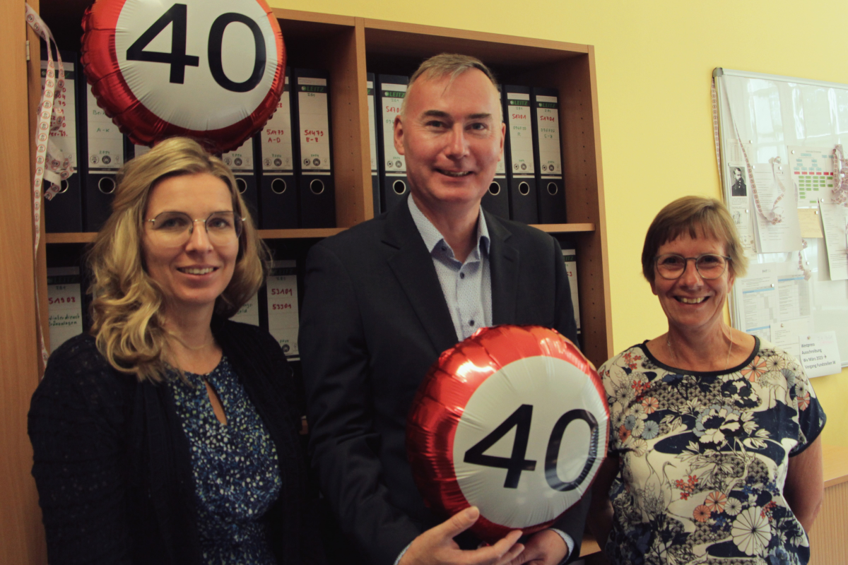 Drei Personen in einem Büro, der Mann in der Mitte hält einen Luftballon mit der Aufschrift „40“.