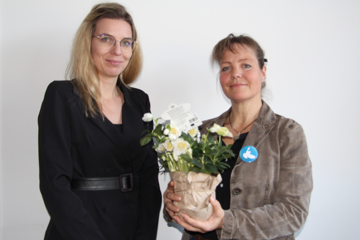 Zwei Frauen stehen vor einer weißen Wand. Eine hält einen Blumenstrauß in der Hand.